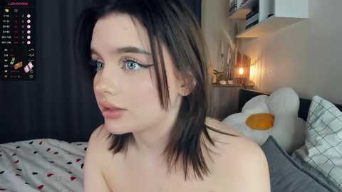 Media: Video of a young Caucasian woman with shoulder-length dark hair, light skin, and blue eyes, topless in a bedroom with a white teddy bear, a TV showing a calendar, and a lamp.