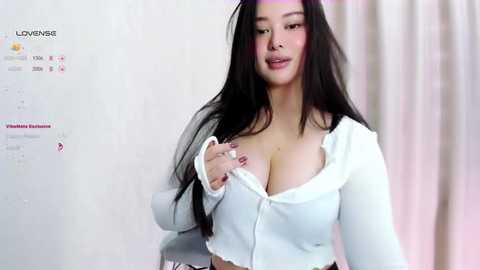 Media: Video of a young woman with long black hair, light skin, and large breasts, wearing a white cropped cardigan, revealing cleavage, standing in front of a white wall with a \"Lovenese\" watermark.