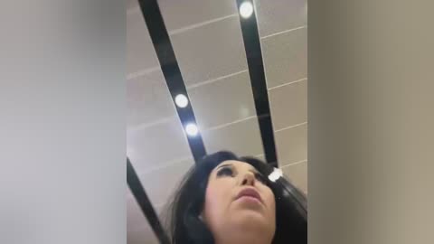 Media: Video of a woman with long black hair, wearing makeup, standing in an office with a tiled ceiling, black ceiling lights, and a blurred background.