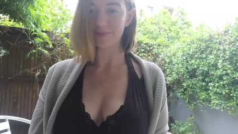 Media: Video of a light-skinned woman with shoulder-length brown hair, wearing a black lace bra and a beige cardigan, standing outdoors against a lush green background with a wooden fence.