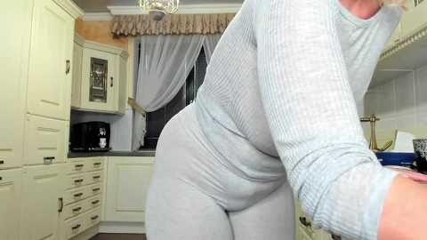 Media: Video of a woman in a light gray ribbed long-sleeve top and matching pants, bending over in a modern kitchen with white cabinets, a black microwave, and a window with sheer curtains.