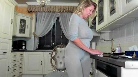 Media: A video of a blonde woman in a light gray, tight-fitting outfit, washing dishes in a modern, white kitchen with tiled backsplash and dark countertops.