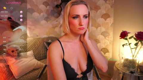 Media: A video of a blonde woman with fair skin, wearing black lace lingerie, sitting in a dimly-lit bedroom with floral wallpaper and rose petals.