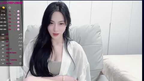 Media: A video of an East Asian woman with long black hair, wearing a white robe, sitting on a white couch. She has fair skin and a slender build.