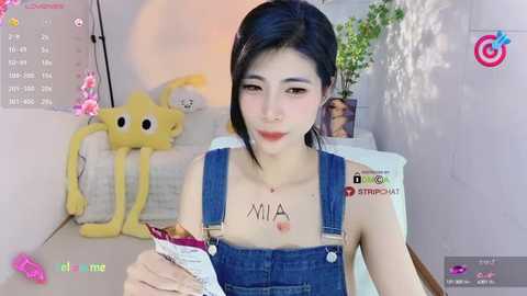 Media: Video of a young East Asian woman with fair skin, dark hair, wearing blue overalls. She's seated indoors, holding a magazine with \"MIA\" written on her chest. Background features a yellow plushie and a potted plant.