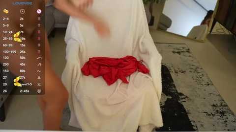 Media: Video of a person in a white robe, holding a red cloth, standing in a modern living room with a patterned rug.