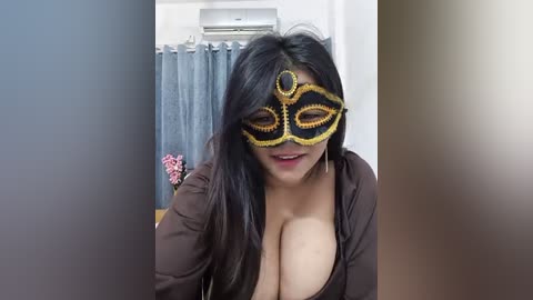 Media: Video of a woman with long black hair, wearing a revealing black and gold masquerade mask, low-cut brown top revealing ample cleavage, indoors with a blue curtain and air conditioning unit in the background.