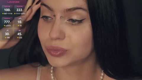 Media: A close-up video of a young woman with fair skin, long black hair, and light makeup, including subtle eyeliner and mascara, holding a makeup brush against her forehead. She wears a pearl necklace.