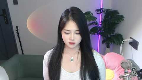 Media: Video of an East Asian woman with long black hair, wearing a light gray top and necklace, in a modern room with a green couch, plants, and purple lighting.