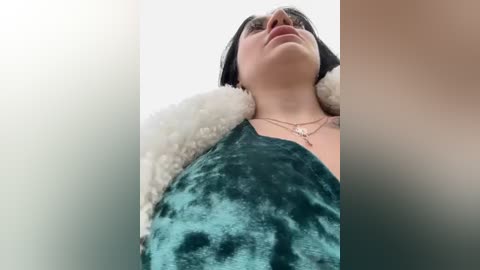 Media: Video of a young woman with fair skin, black hair, and a teal velvet dress, wearing a white fur coat and silver necklace, standing against a blurred background.