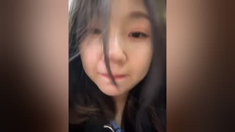 Media: A blurry video of a young Asian woman with long, dark hair, wearing a dark jacket, and a soft smile, taken indoors with a brown background.