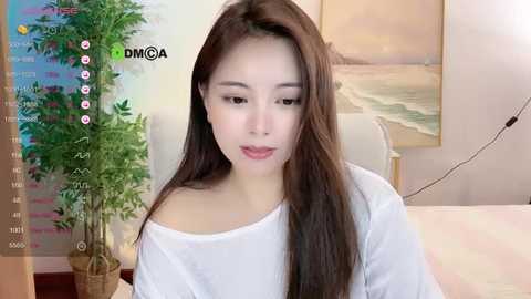 Media: Video of a young East Asian woman with long brown hair, wearing a white off-shoulder top, sitting indoors with a potted plant and framed beach painting behind her.