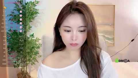 Media: Video of an East Asian woman with long, straight brown hair, wearing a white off-shoulder top, in a modern room with a bamboo plant, wall art, and a desk lamp.