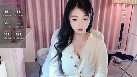 Media: Video of an East Asian woman with long black hair, fair skin, and large breasts, wearing a white blouse, sitting indoors with pink curtains and a lamp.