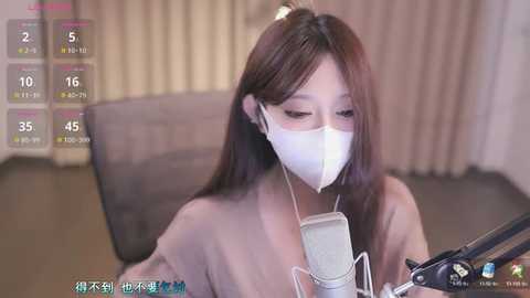 Media: Video of an Asian woman with long brown hair, wearing a white face mask, headphones, and a beige sweater, recording a podcast on a microphone.