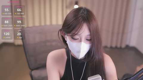 Media: Video of an East Asian woman with long dark hair, wearing a white mask, black top, and headphones, seated on a grey couch in a neutral-toned room.