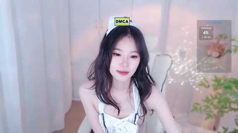 Media: Video of an Asian woman with long black hair, wearing a white maid outfit with a hat reading \"DMCA.\" She sits in a softly lit room with a floral arrangement in the background.