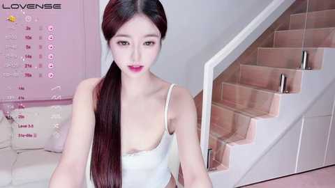 Media: Video of a young Asian woman with long, straight, dark hair in a white tank top, sitting on a beige sofa in a modern, pink-themed room with a staircase.