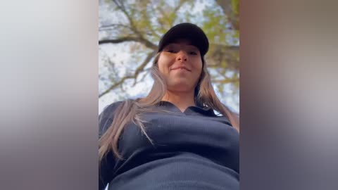 Media: A video of a woman with long, straight, light brown hair, wearing a black cap and a dark gray long-sleeve shirt, smiling downwards. The background shows a blurred tree with green leaves.