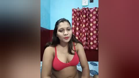 Media: Video of a young Latina woman with medium skin tone, wearing a red bra, sitting on a bed with red and blue patterned sheets. She has long, straight dark hair and is looking directly at the camera. The background includes a red curtain with floral patterns and a blue wall.