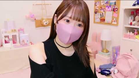 Media: A video of an East Asian woman with straight brown hair, wearing a black off-shoulder top, purple face mask, and a black choker. The background features a pink and white-themed bedroom with various decorations and a bulletin board.