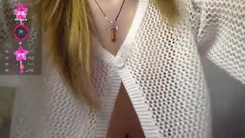 Media: Video of a young woman with long blonde hair wearing a white, textured cardigan, revealing a pink bra underneath. She has a necklace with an amber pendant. The background is blurred, featuring a pink star icon.