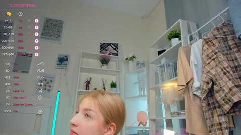 Media: A video captures a young woman with blonde hair, wearing a beige jacket, standing in a minimalist, white-walled room with a calendar, potted plants, and a white shelf filled with books and clothes.