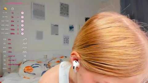 Media: Video of a blonde woman wearing white headphones, lying on a bed with colorful bedding. The room has a minimalistic decor with a few posters and a white wall.