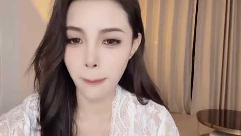 Media: A video of an East Asian woman with long, straight black hair, fair skin, and delicate facial features. She wears a light-colored, lace-trimmed robe, standing in front of beige curtains and a round wooden table.