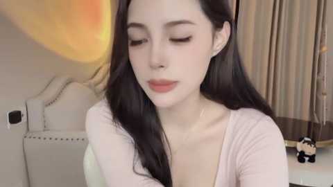 Media: A video of a young East Asian woman with long, straight black hair, fair skin, and closed eyes, wearing a light pink top. She sits indoors next to a white chair with a round cushion.