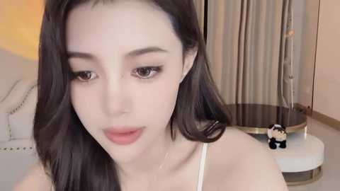 Media: A close-up video of an Asian woman with fair skin, long black hair, and full lips. She wears a white spaghetti-strap top. Background shows a modern bedroom with beige curtains and a plush chair.