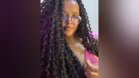 Media: Video of a young Black woman with medium brown skin, large round glasses, and voluminous black curly hair, smiling while touching her hair. She wears a pink top.