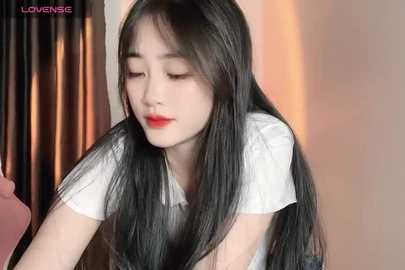 Media: Video of a young Asian woman with long, straight black hair and fair skin, wearing a white blouse, leaning forward, looking down, in a softly lit room with beige walls and a pink curtain.