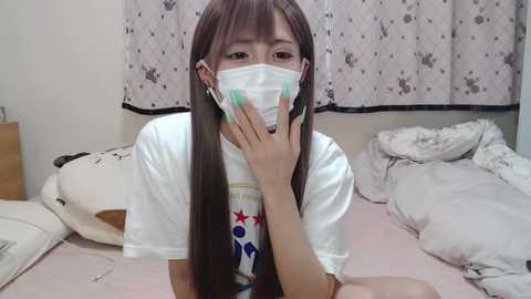 Media: Video of a young Asian woman with long brown hair, wearing a white T-shirt, face mask, and earrings, sitting on a bed with messy white sheets, surrounded by stuffed animals.