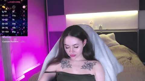 Media: Video of a young woman with long brown hair, wearing a dark green strapless dress, standing in a modern bedroom with a bed, a calendar display, and purple LED lights.