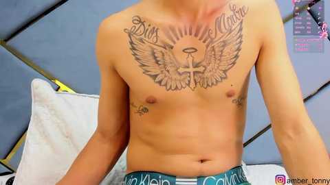 Media: Video of a shirtless, light-skinned male with tattoos including a large angel wings design and \"Eternal Love\" across his chest. He wears blue Calvin Klein underwear. Background shows a blue bed with white pillows and a pink poster.