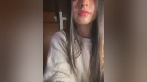 Media: Video of a young woman with straight brown hair, fair skin, and full red lips, wearing a white sweater, taken indoors with soft lighting.