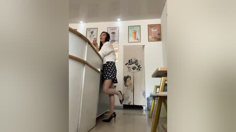 Media: Video of an Asian woman with medium build, short black hair, wearing a white blouse, black polka-dot skirt, and high heels, leaning on a white countertop in a modern, minimalist kitchen.