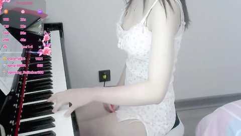 Media: Video of a slender, fair-skinned woman with long dark hair, wearing a white lace lingerie bodysuit, playing a black upright piano in a dimly lit room.