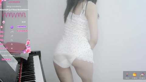 Media: Video of a young Asian woman with long black hair, wearing a white lace camisole and matching shorts, standing in front of a piano. Background includes digital text and images.