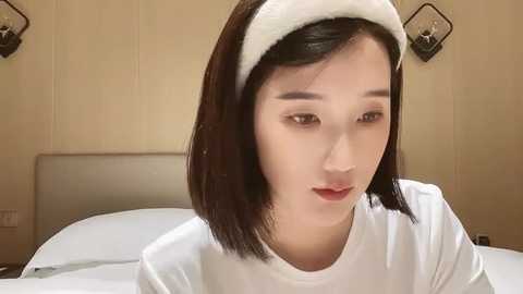 Media: A video of an East Asian woman with straight black hair, fair skin, wearing a white headband and white shirt, sitting on a bed in a beige bedroom.