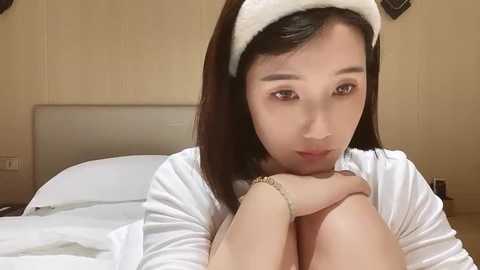 Media: A video of an Asian woman with shoulder-length black hair and a white headband, wearing a white long-sleeved top, sitting on a bed with white sheets, looking contemplative.