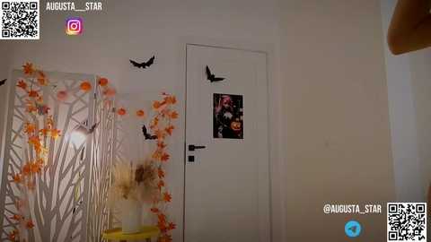 Media: A video of a modern, minimalist room with a white door adorned with a Halloween-themed sticker and black bats. The walls are decorated with orange and white foliage patterns. QR codes and social media handles are visible.