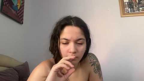 Media: Video of a young woman with dark hair, fair skin, and a tattooed arm, biting her finger, leaning on a couch against a wall with colorful art and a framed collage.