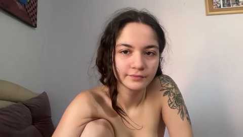 Media: Video of a young, light-skinned woman with long, wavy dark hair, wearing no clothes, revealing a tattoo on her upper arm. She sits on a bed with beige sheets, against a light wall with framed art.