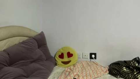 Media: A video of a simple bedroom corner featuring a yellow emoji with red heart eyes, a lavender pillow, and a beige quilt.