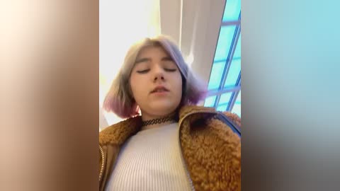 Media: Video of a young woman with light skin and shoulder-length, pastel pink hair. She wears a ribbed cream sweater and a brown fuzzy jacket. Background includes a window with vertical blinds and soft lighting.