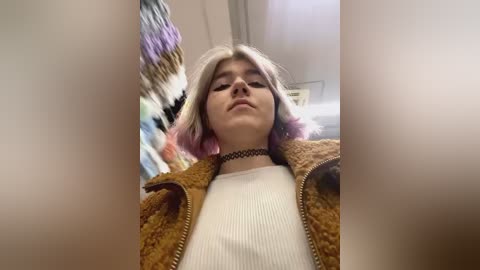 Media: Video of a young woman with pastel-colored hair, wearing a choker and a brown knit jacket over a white ribbed sweater, standing in a brightly lit, colorful room.