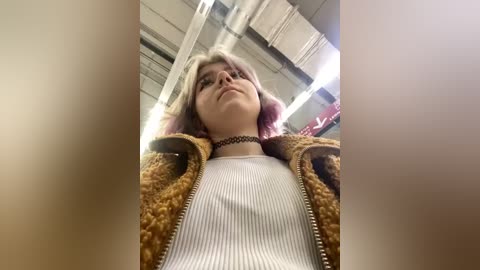 Media: A video of a young woman with short, dyed pink hair and fair skin, wearing a choker, striped shirt, and brown jacket, standing in a brightly lit, industrial setting with exposed pipes and ceiling lights.