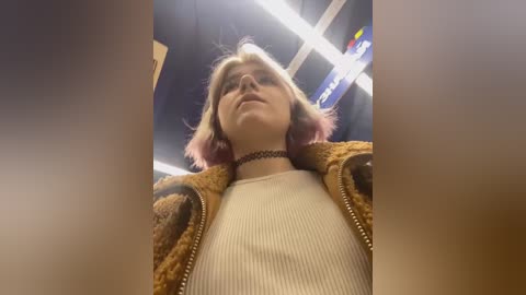 Media: Video of a young woman with short, pink-tinted hair, wearing a beige ribbed sweater and a brown, patterned jacket, standing in a dimly lit, modern interior.